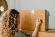 Back view of unrecognizable curvy female in casual clothes writing fragile on empty side of carton box while waiting for courier