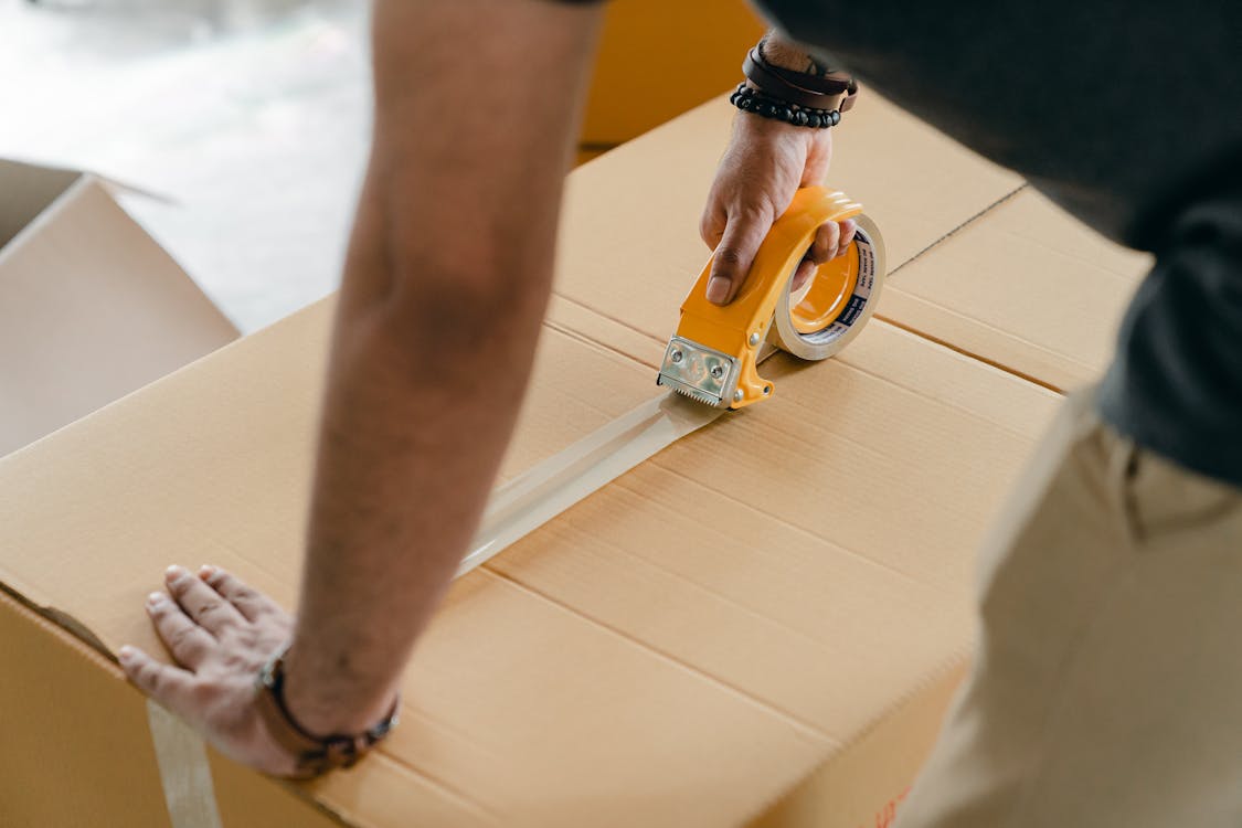 Crop faceless man packing box with scotch tape