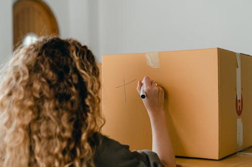 Back view of faceless young lady with curly hair preparing cardboard boxes for moving