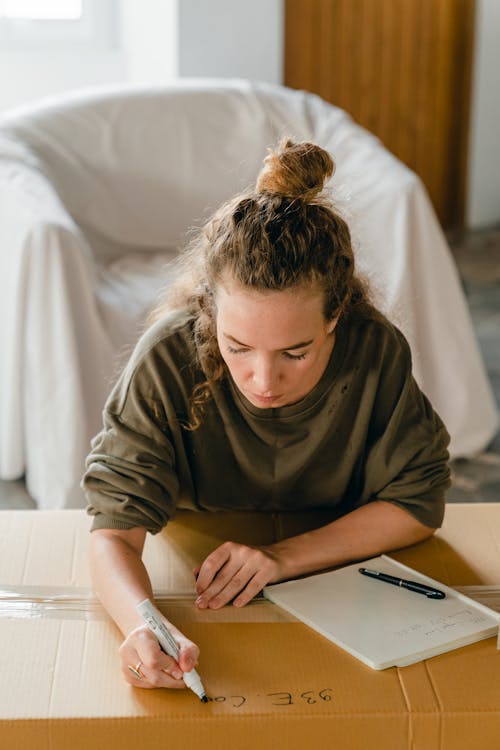Free From above of concentrated young woman checking and taking notes on cardboard box while preparing for relocation Stock Photo