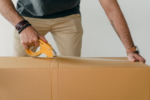 Free Crop anonymous young male packer in casual clothes using tape gun dispenser for sealing cardboard boxes Stock Photo