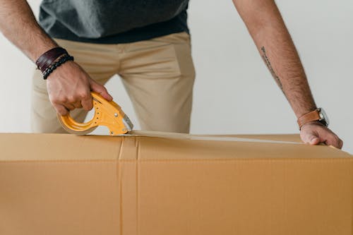 Crop anonymous young male packer in casual clothes using tape gun dispenser for sealing cardboard boxes