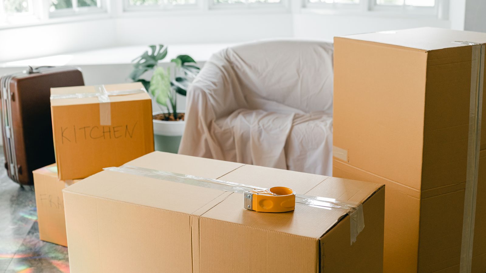 Cardboard boxes and a suitcase stacked on the floor of a bright room before a move, a real stock photo with no people in the frame that sets the scene for Nobody Wants This Season 3, where Joanne and Noah move in together