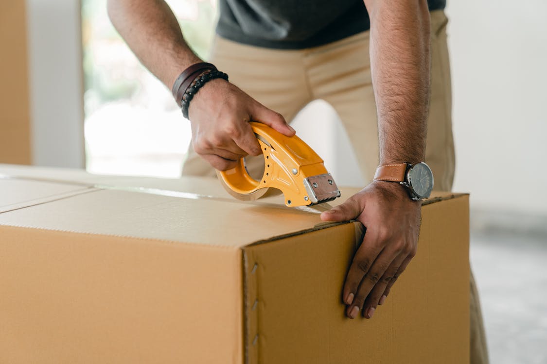 Free Crop Faceless Young Male With Wristwatch Using Adhesive Tape While Preparing Cardboard Box For Transportation Stock Photo