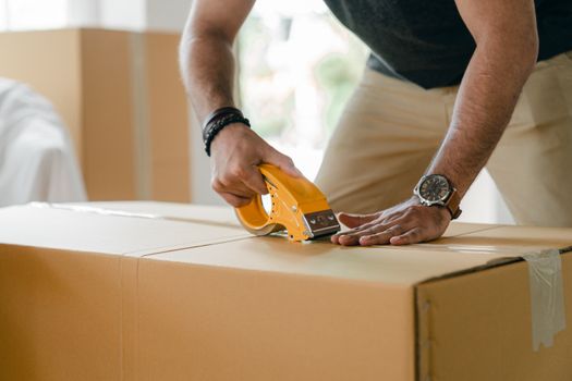 Crop unrecognizable young male in casual clothes packing cardboard box and sealing with scotch tape