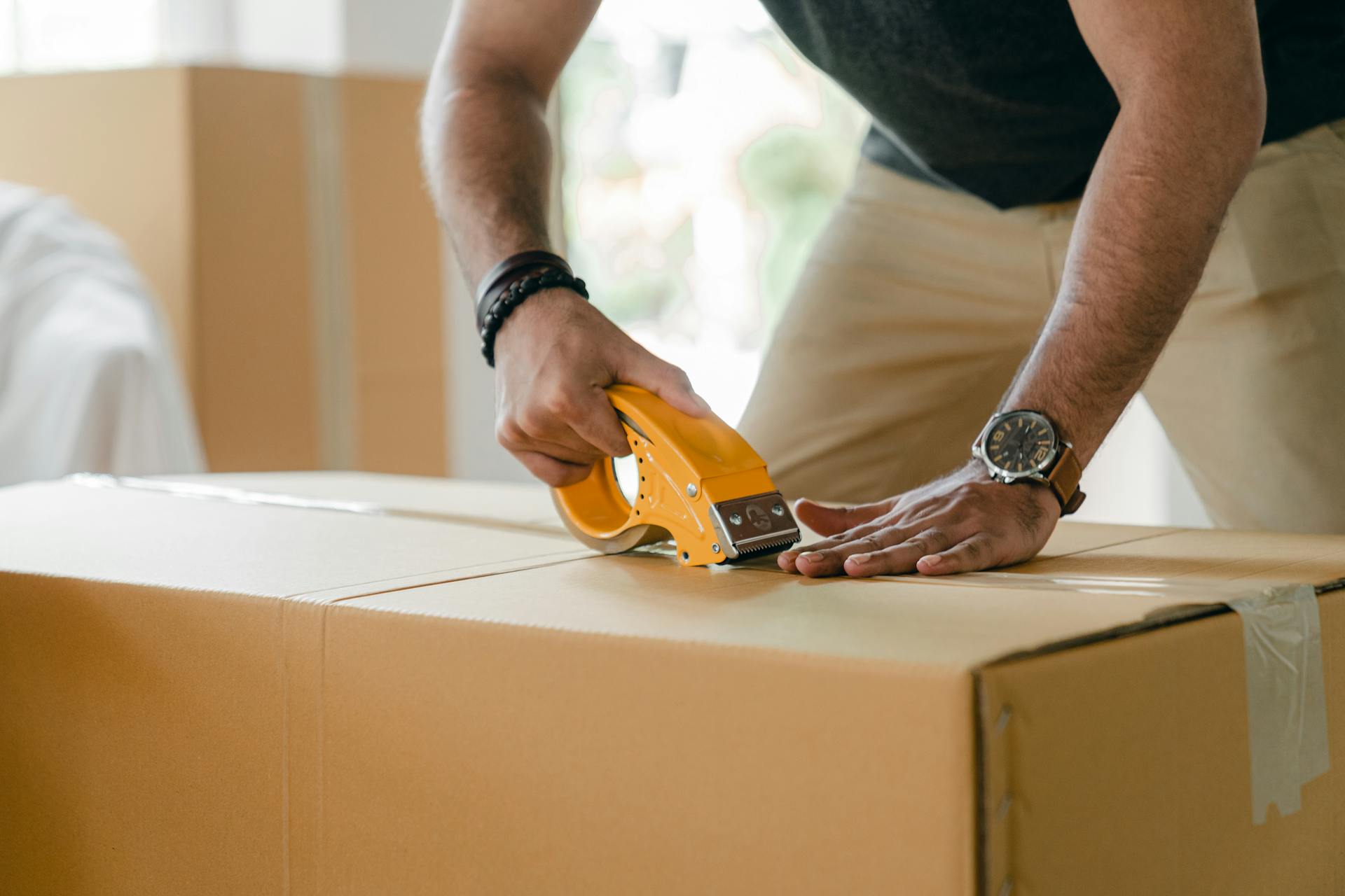 Crop unrecognizable young male in casual clothes packing cardboard box and sealing with scotch tape