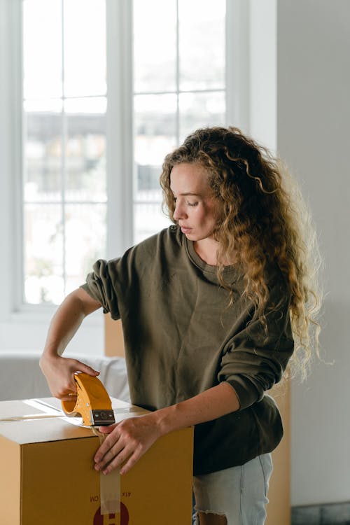 Young concentrated woman taping boxes using packing instrument