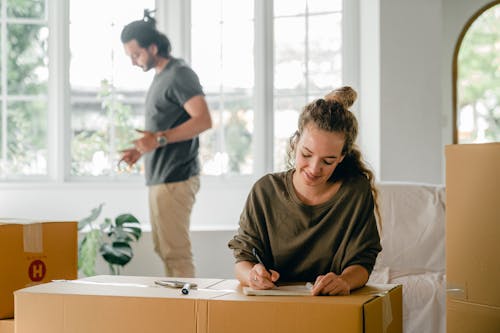 Positive young woman writing in notepad while sitting against cardboard boxes during relocation in new house with husband