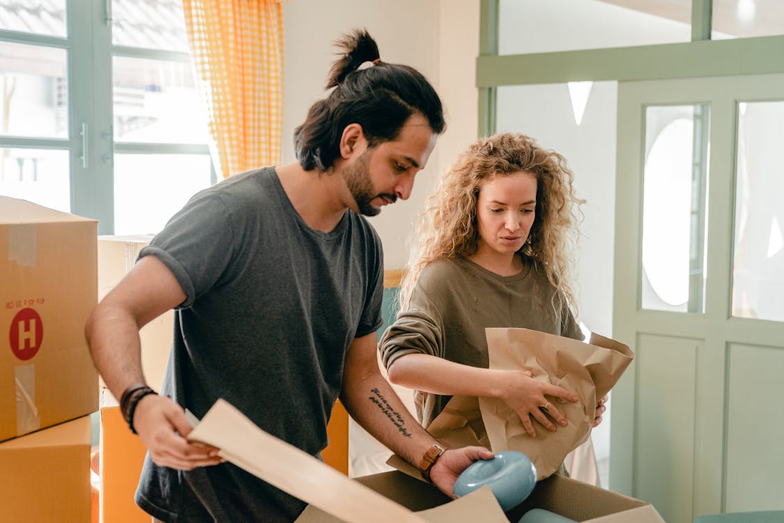 Concentrated young diverse couple using wrapping paper while packing belongings in carton boxes during relocation