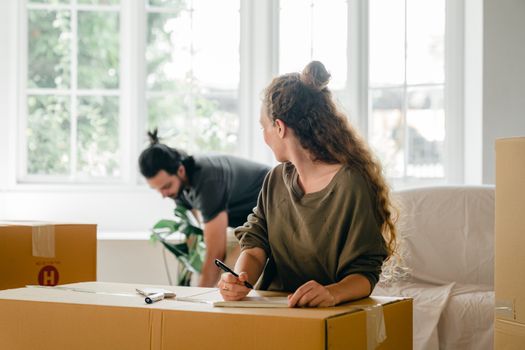 Unrecognizable woman with pen and notebook sitting near heap of cardboard boxes and looking at ethnic boyfriend with ponytail standing near large window in apartment