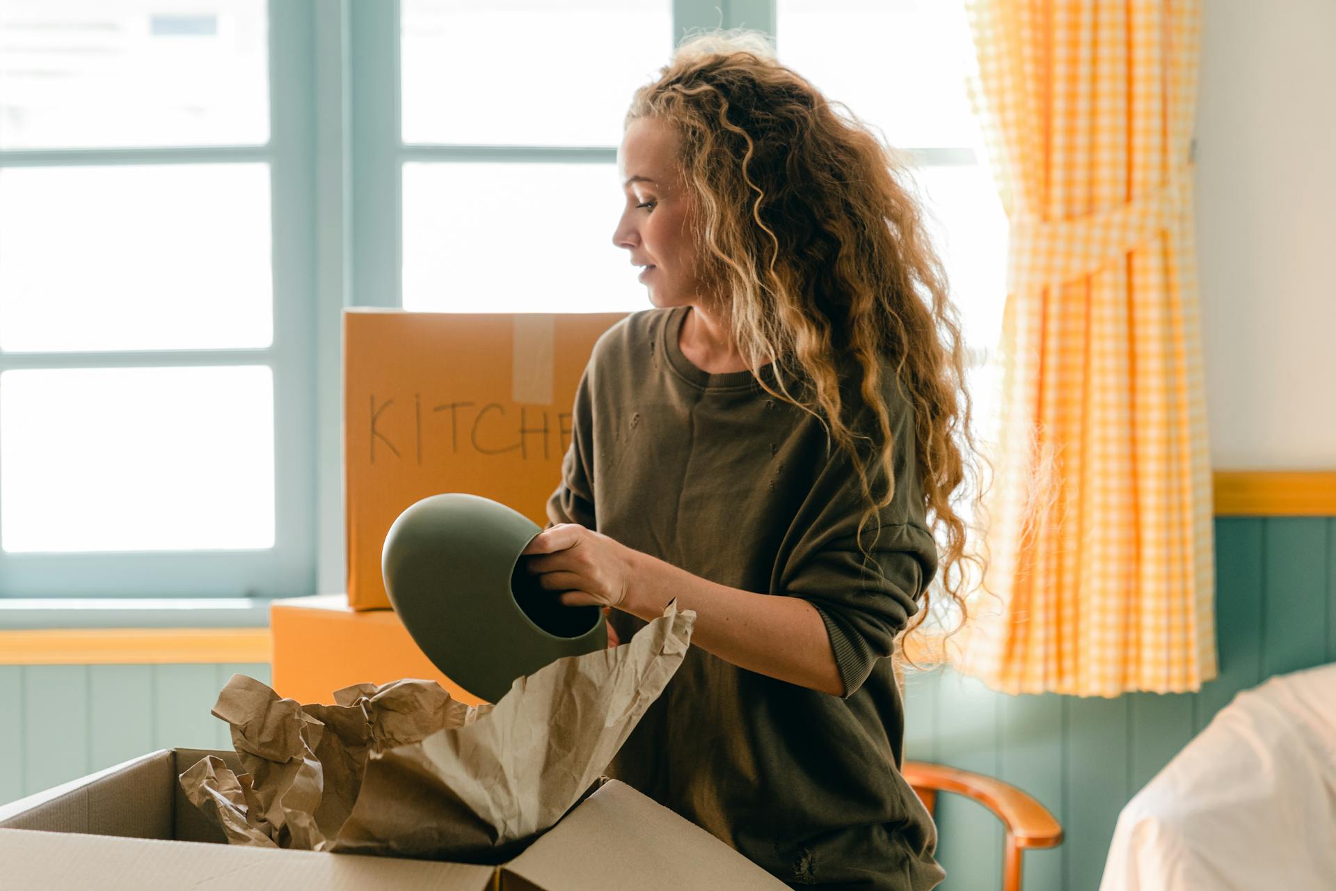 Dreamy female in casual wear taking ceramic vase out of parchment while standing near cardboard box with KITCHEN inscription in back lit preparing to move into new house