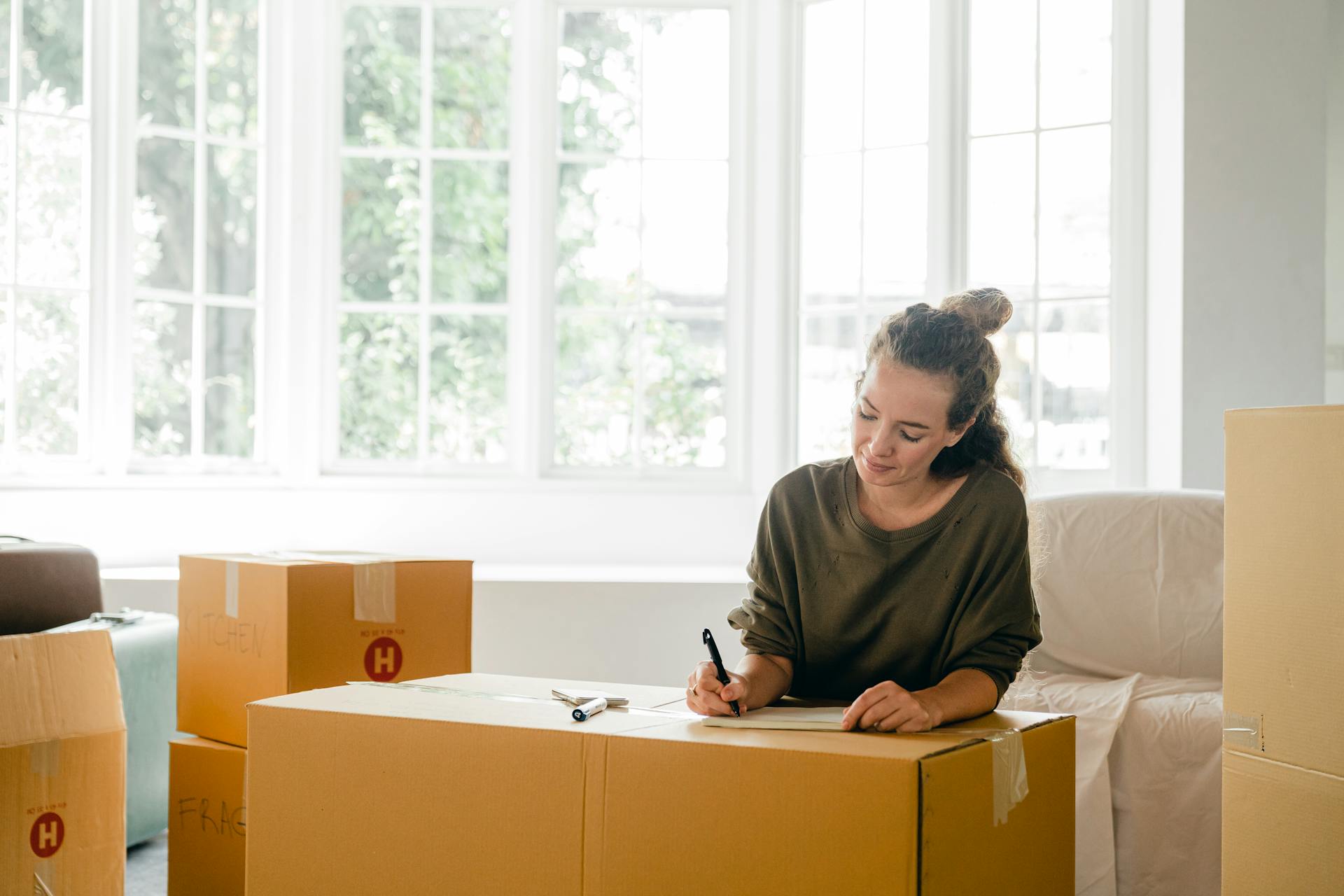 Content lady in casual clothes writing with pen on cardboard box while sitting near window in daylight in apartment preparing to relocate