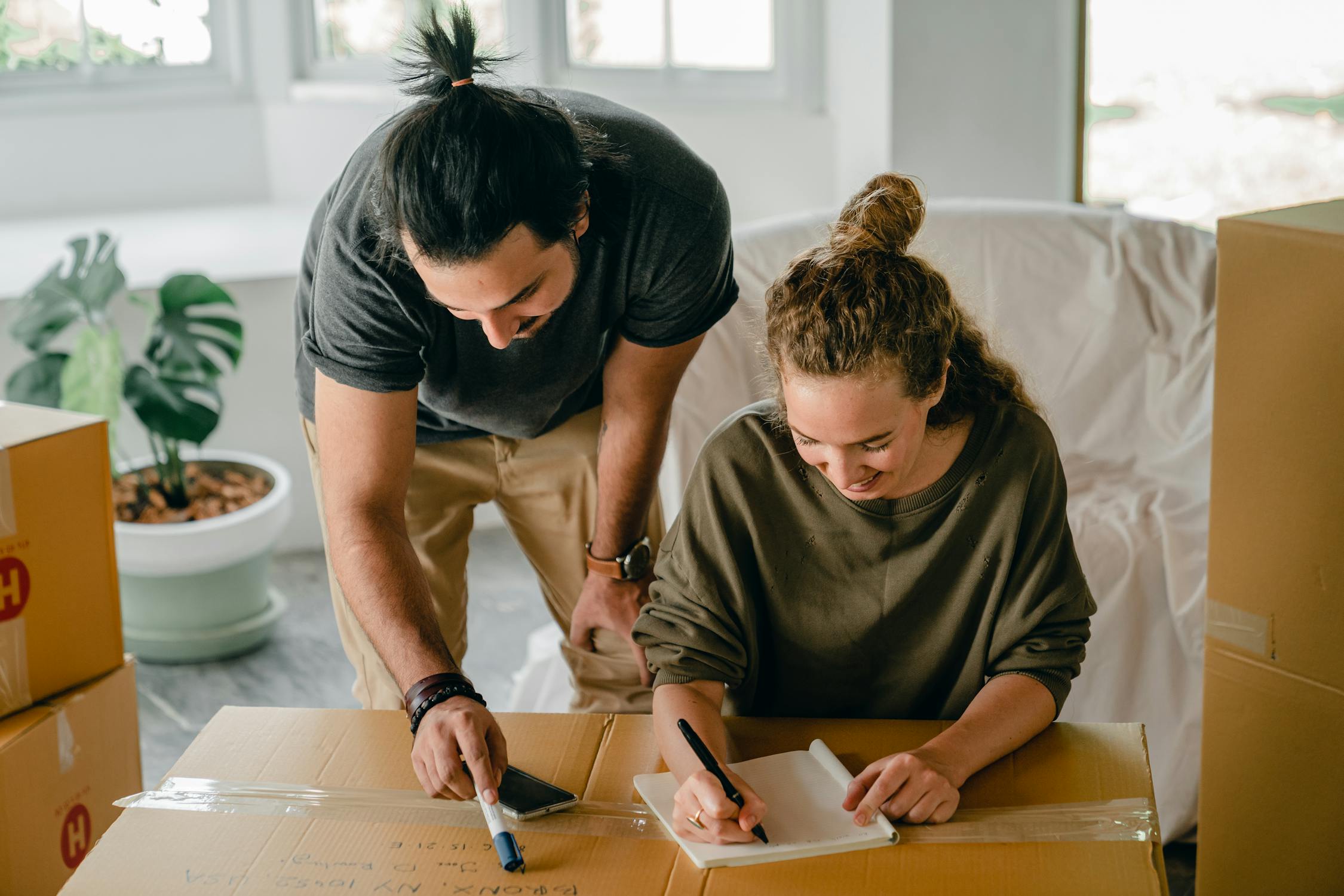 Cheerful diverse couple writing in notebook near boxes before ...
