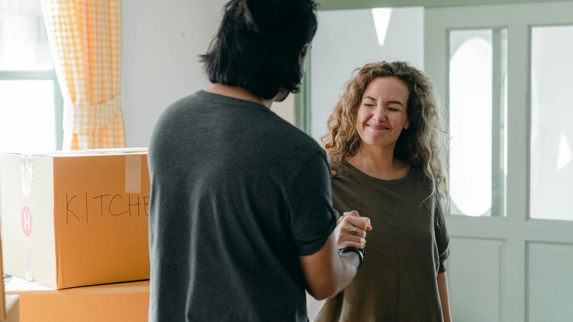 Cheerful girlfriend with closed eyes shaking hand to crop unrecognizable male partner in casual wear while standing near box with KITCHEN inscription after moving into new flat