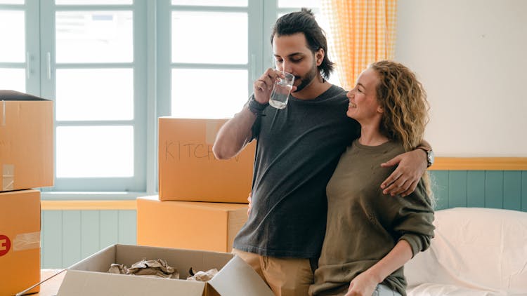 Cheerful Multiethnic Couple Embracing While Drinking Water Near Different Boxes