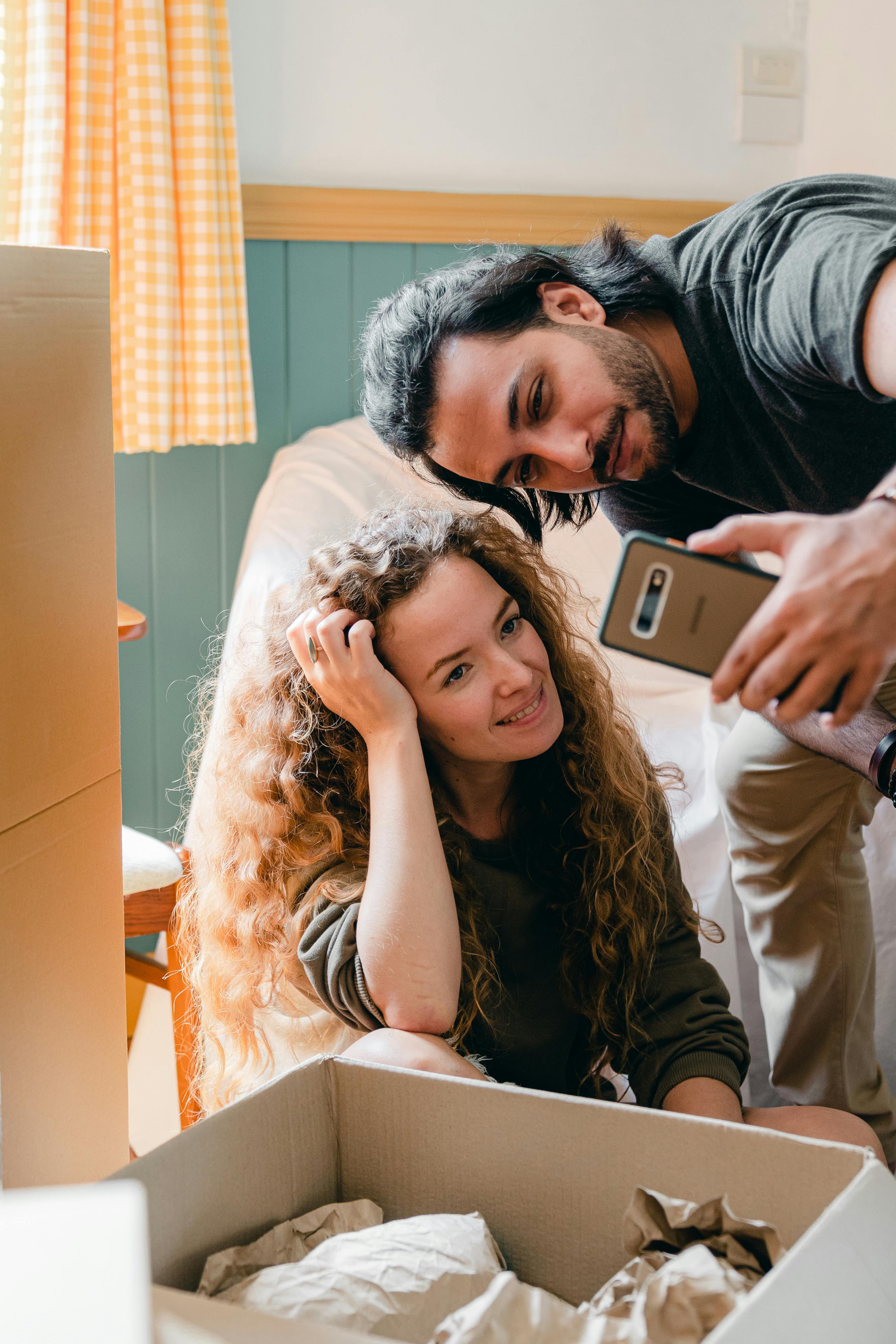 multiracial couple taking selfie on smartphone while unpacking in new home