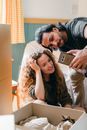 Happy girlfriend with wavy hair and cheerful ethnic boyfriend taking selfie on cellphone while standing in back lit near heap of cardboard boxes after moving into new apartment
