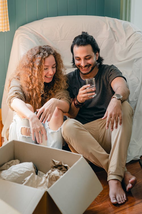 Free Happy ethnic barefoot man sitting with glass of water on comfortable sofa while talking to positive girlfriend near open cardboard box with parchment at home Stock Photo