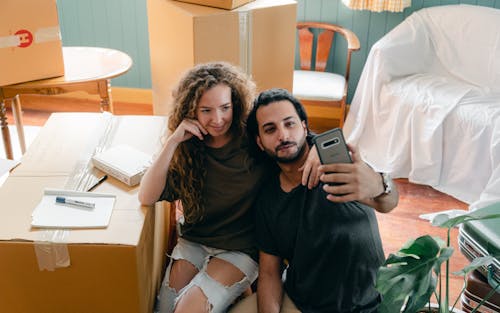 From above of positive young diverse couple in casual clothes bonding and taking selfie on modern smartphone while resting after packing belongings in carton boxes to relocate