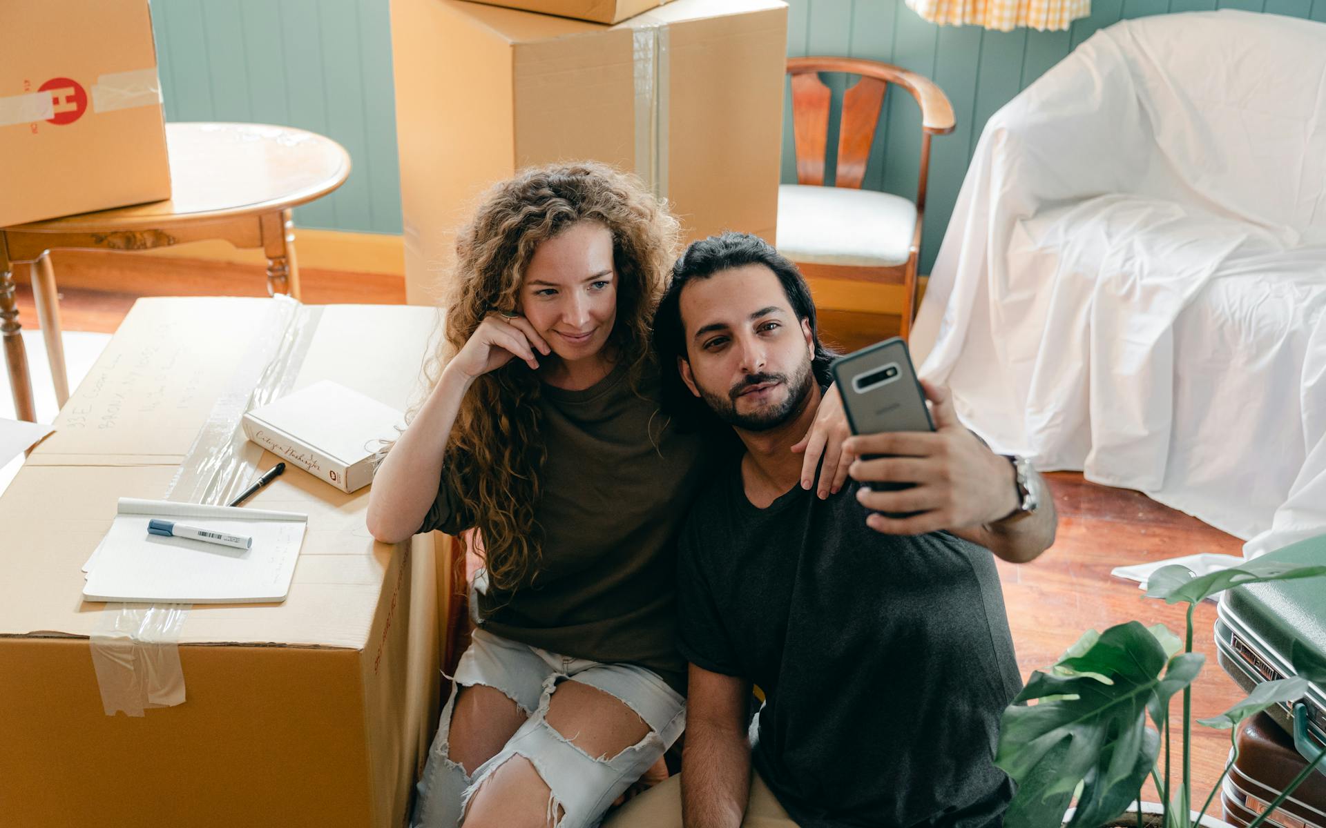 From above of positive young diverse couple in casual clothes bonding and taking selfie on modern smartphone while resting after packing belongings in carton boxes to relocate
