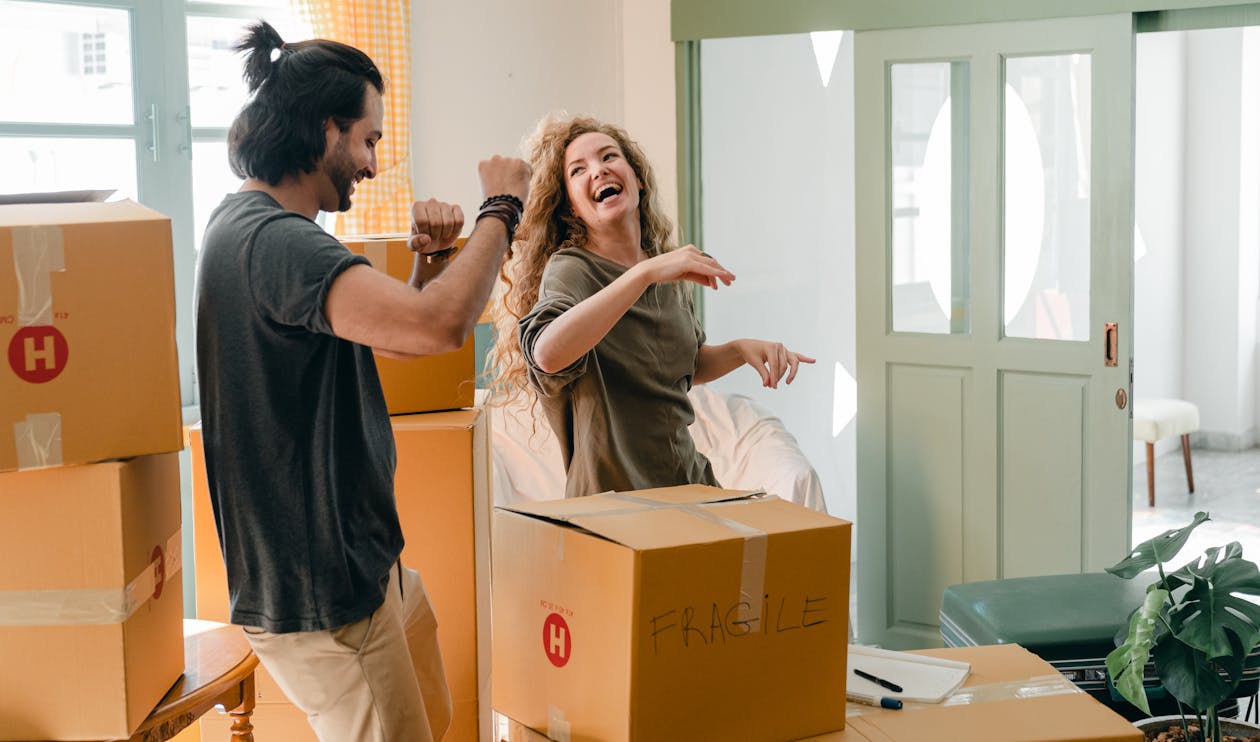 Cheerful laughing couple in casual clothes having fun and dancing together while unpacking carton boxes after moving into new contemporary apartment