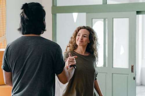 Cheerful young couple in casual outfits smiling and shaking hands while spending time together in light modern apartment and looking at each other happily