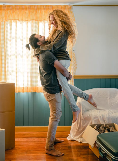 Full body barefoot bearded happy man in casual clothes lifting young smiling girlfriend up while moving together into new cozy apartment