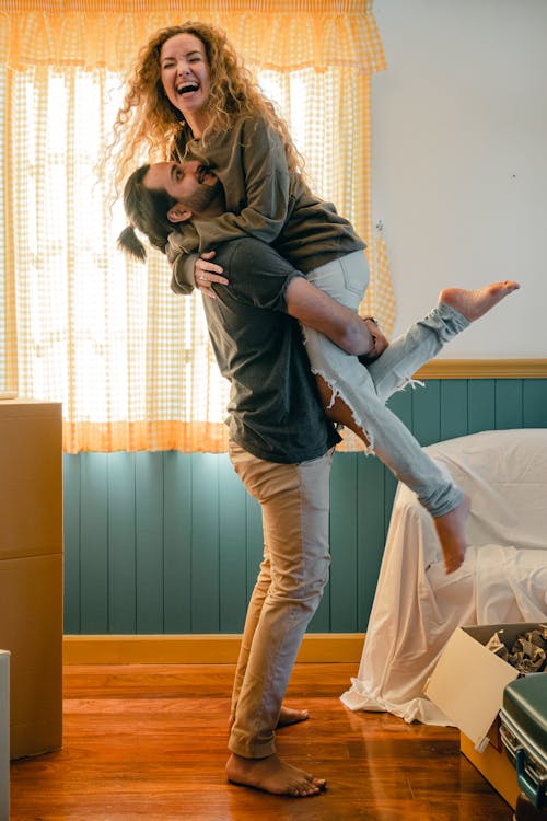 Free Side view full body barefoot happy bearded male in casual wear lifting cheerful laughing girlfriend up while unpacking belongings from carton boxes in new cozy apartment Stock Photo