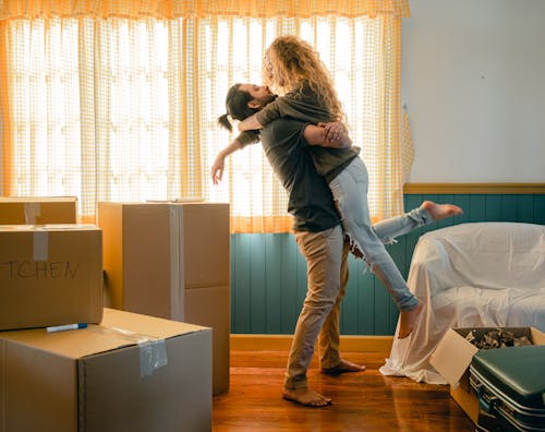 Free Side view full body barefoot young man lifting girlfriend in casual wear with curly hair while unpacking belongings from cardboard boxes and suitcases in light living room Stock Photo