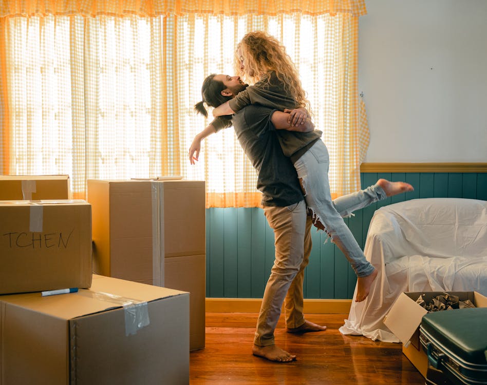Side view full body barefoot young man lifting girlfriend in casual wear with curly hair while unpacking belongings from cardboard boxes and suitcases in light living room