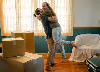 Side view full body barefoot young bearded male in casual clothes standing against window and lifting laughing girlfriend up during relocation and unpacking things from cardboard boxes
