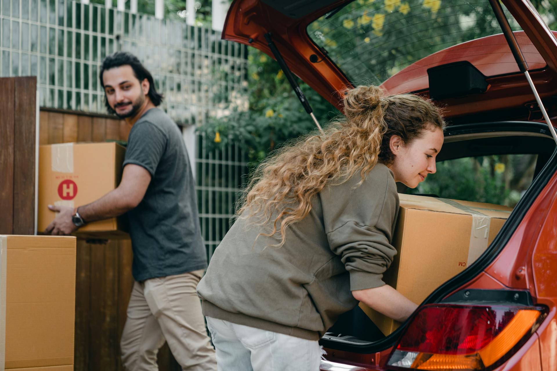 Positive multiethnic boyfriend and girlfriend unloading car trunk during moving to new home in quiet green yard on summer day