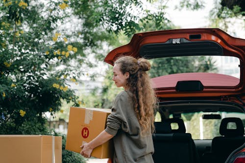 Positive young woman in casual sweatshirt taking cardboard boxes out of trunk compartment while moving into new home with blossoming garden
