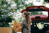 一位穿着休闲运动衫的年轻女子正从后备箱取出纸板箱，准备搬进花园盛开的新家