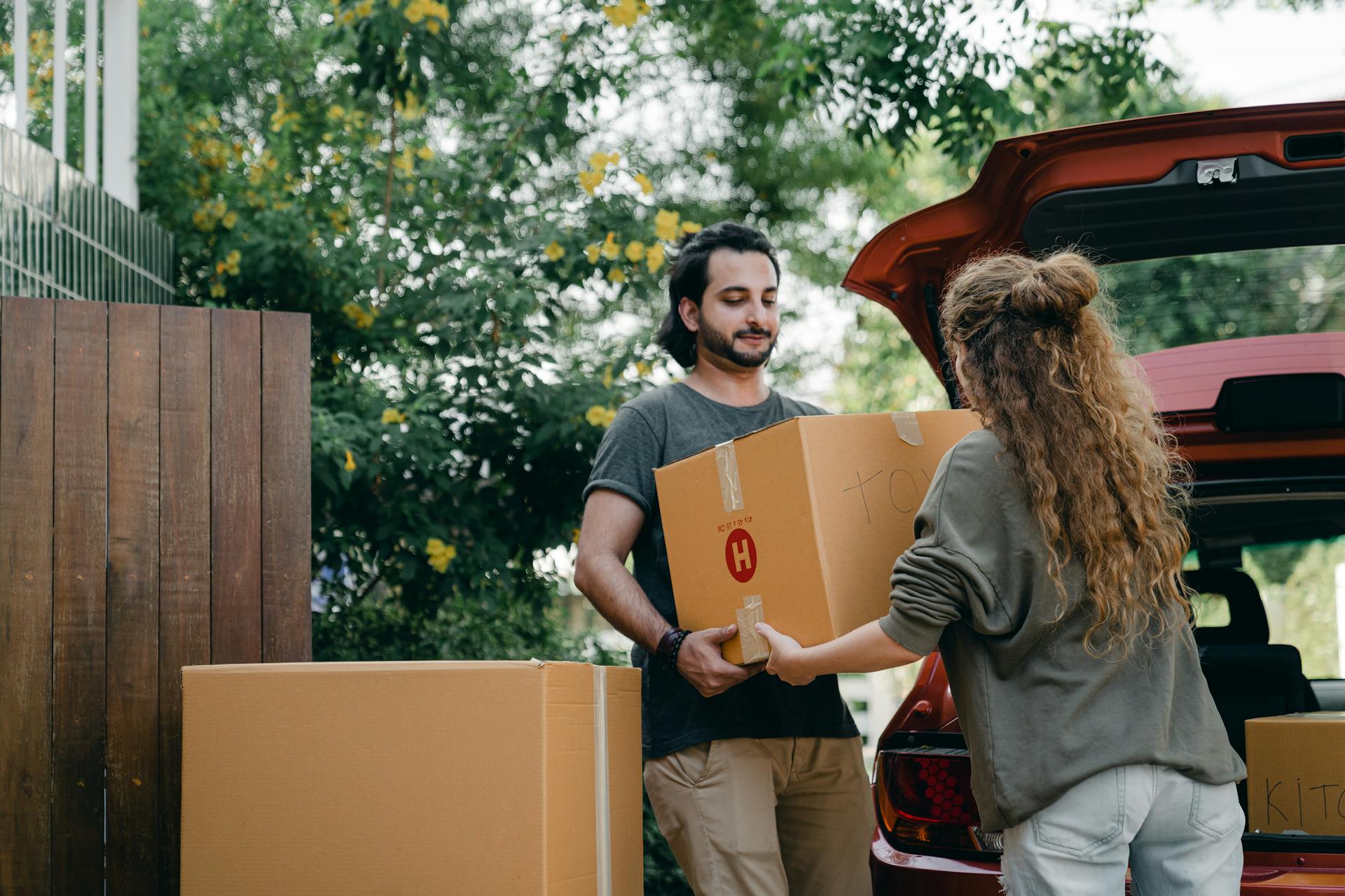 Boyfriend and girlfriend in casual wear helping each other with unpacking car while moving in together on sunny summer day