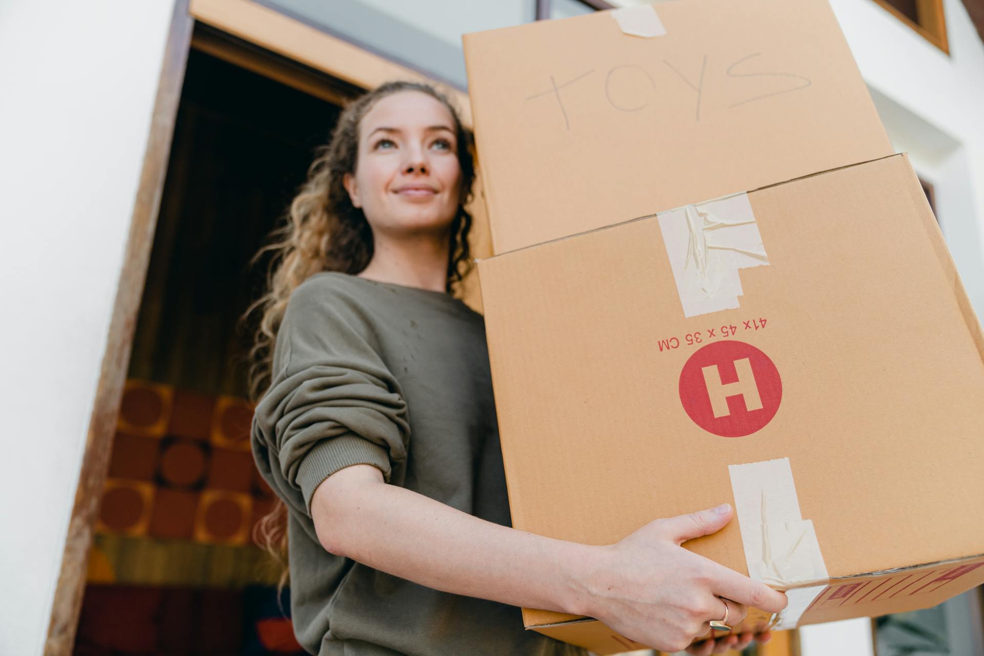 Confident young lady in casual wear carrying heavy carton boxes with toys and looking away while leaving old house