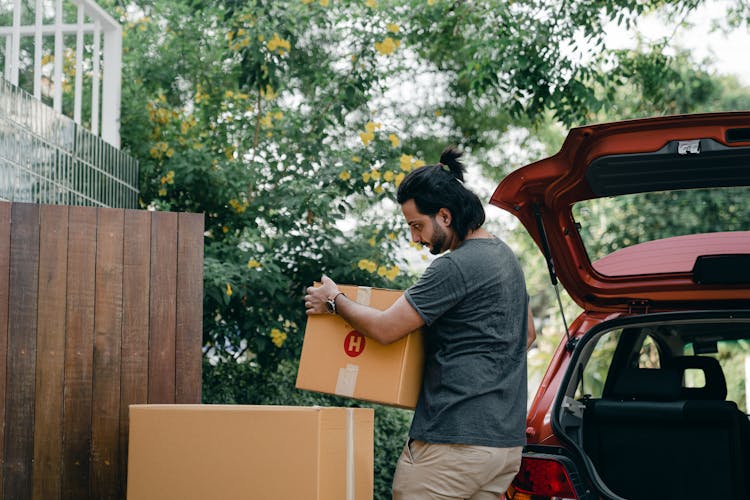 Young Male Taking Boxes Out Of Luggage Boot Of Car