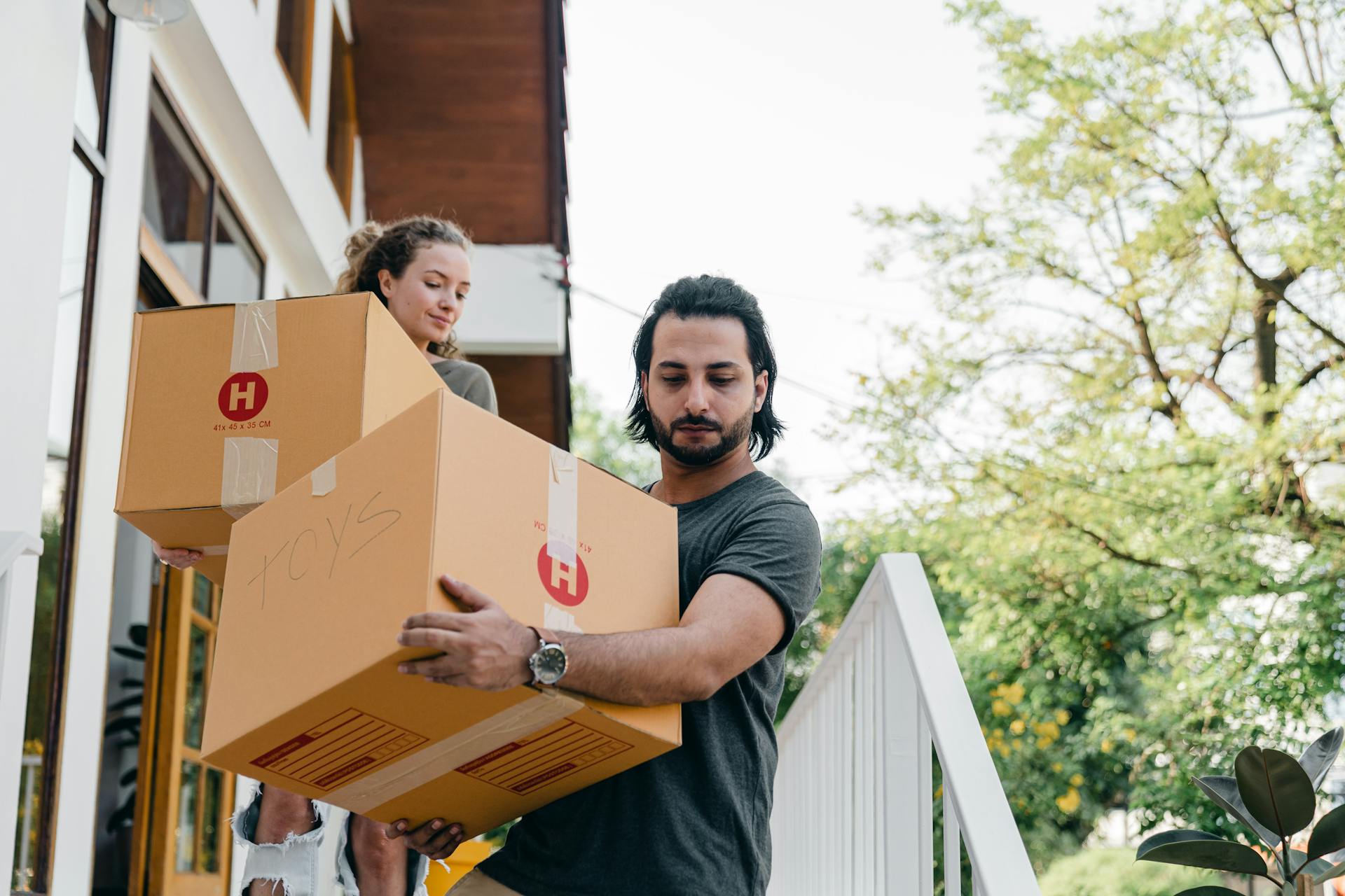 Young multiracial boyfriend and girlfriend in casual wear carrying big carton boxes and watching step on porch stairs while moving out of old home on sunny day in summer