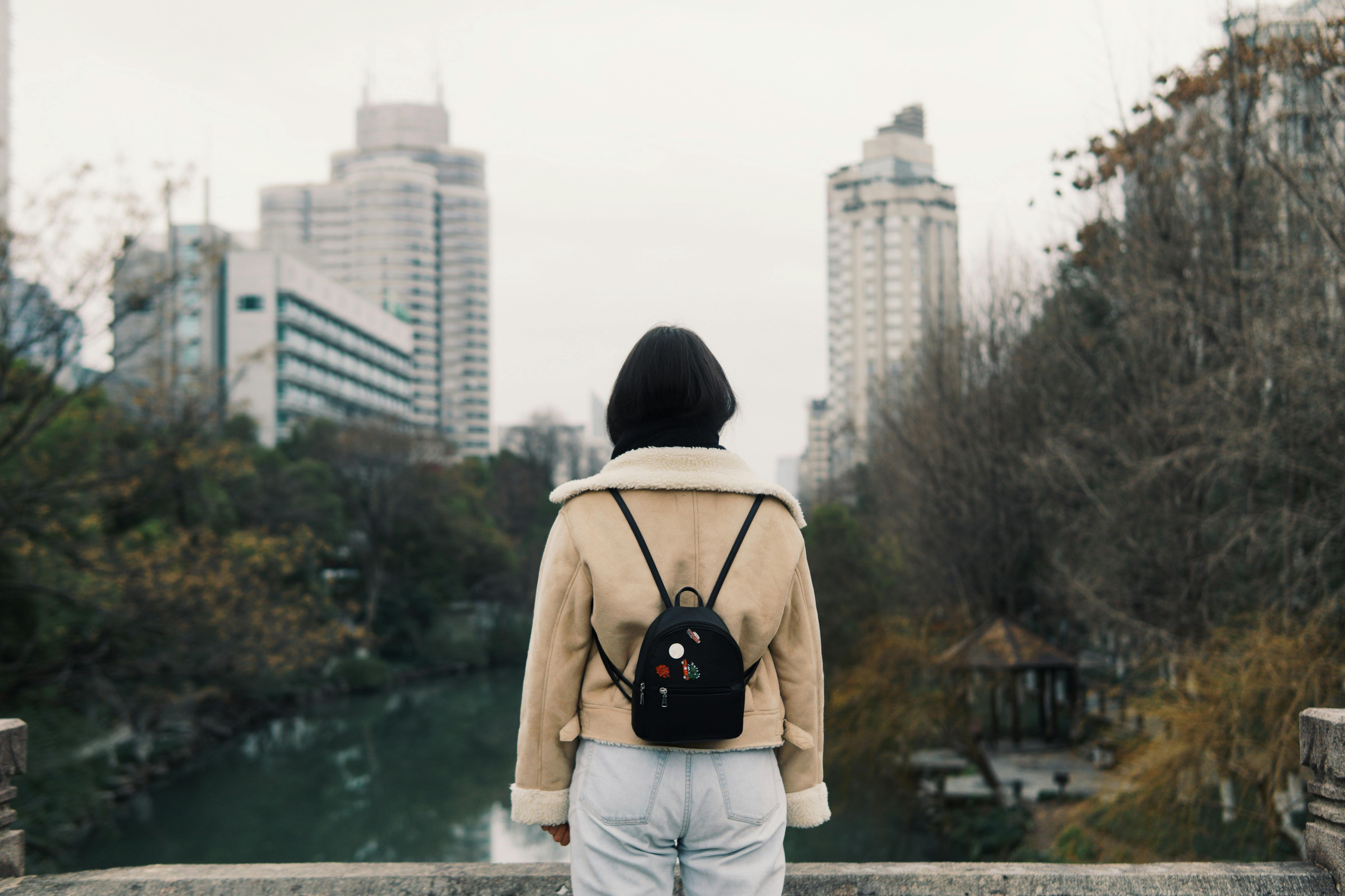 Back view of anonymous female in outerwear with small leather backpack standing on bridge over river between autumn trees with multistory house facades far away in town under sky
