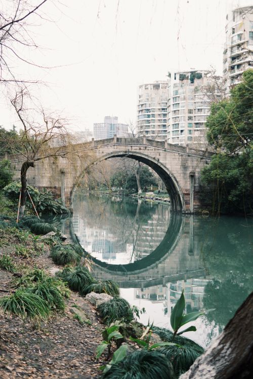 Free A serene view of a stone bridge over a river flanked by urban buildings and lush greenery. Stock Photo