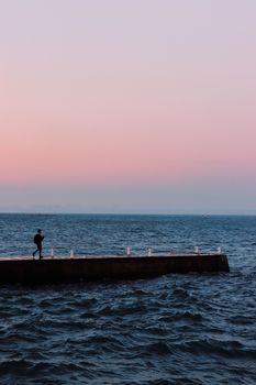 Back view of anonymous person strolling on old stone pier in wavy sea with rippled water and horizon line under sky at sundown in twilight