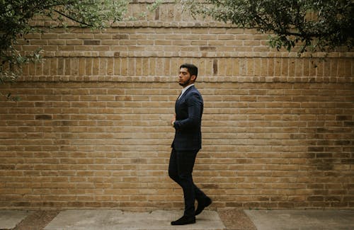 Stylish ethnic man in suit near brick wall in town