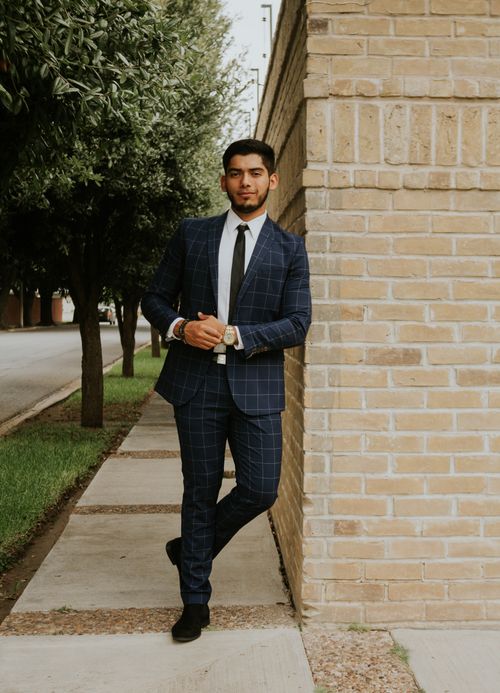 Free Content handsome bearded ethnic man in formal outfit with tie standing on narrow pavement leaned on brick wall near grass with growing trees and looking at camera in town Stock Photo