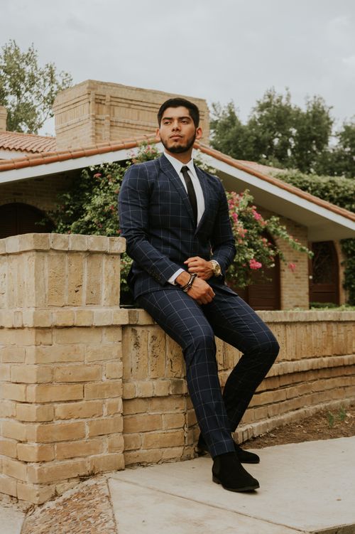 Free Stylish pensive ethnic guy in elegant outfit with tie sitting on old brick fence behind shrub with blooming flowers under sky on city street and looking away Stock Photo