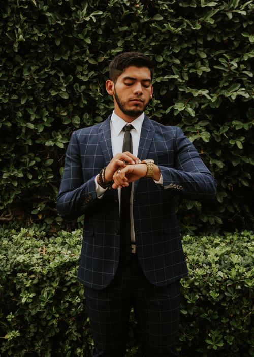 Free Serious ethnic brunet in formal wear with tie looking at wristwatch while standing behind shrubs in daylight Stock Photo