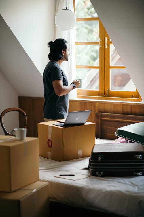 Free Side view of young ethnic male owner of newly bought house with cup of coffee in bedroom full of boxes and suitcases standing and looking at neighboring houses through window Stock Photo