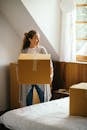 Young smiling lady in casual outfit with big cardboard box in hands standing and looking away near bed in modern bedroom of newly purchased house