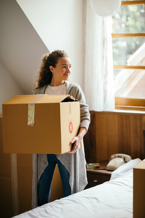 Satisfied female with curly hair smiling while carrying carton box moving out house