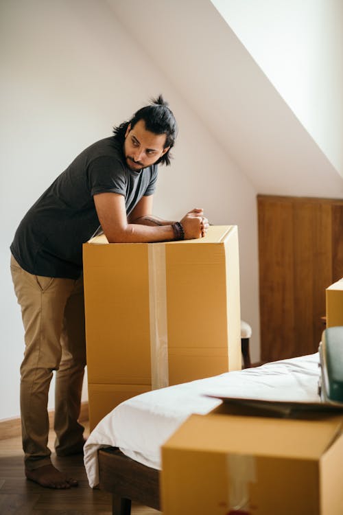 Side view of full body barefoot ethnic man in casual clothes leaning on big cardboard box with stuff from home for moving and looking away