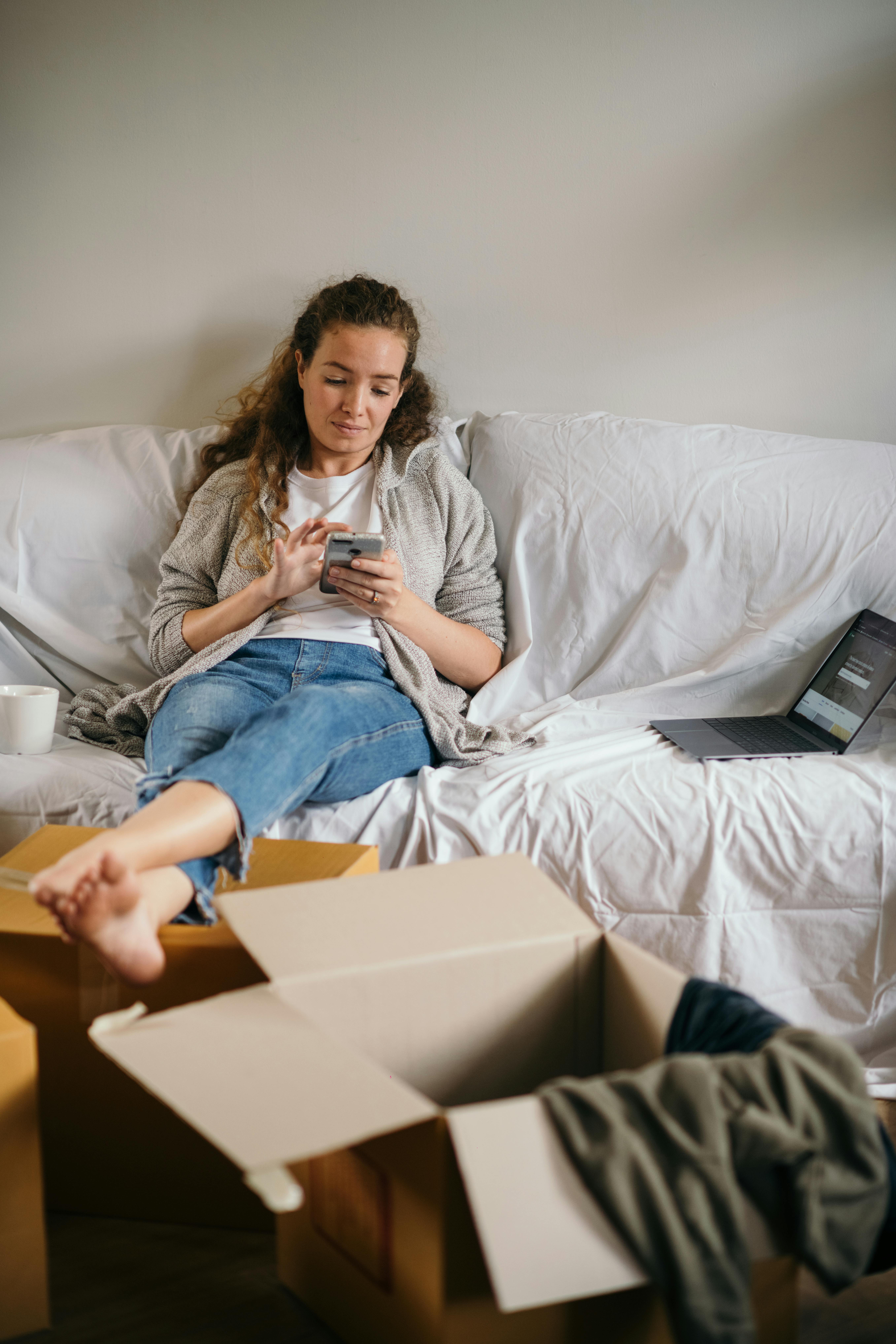 young woman sitting on couch and using smartphone in new house with cardboard boxes